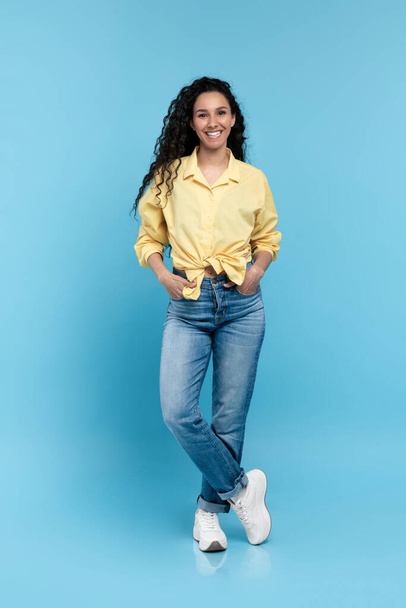 Full length portrait of positive young woman in trendy casual outfit standing and looking at camera over blue studio background. Happy millennial lady in shirt, jeans and sneakers smiling and posing - Photo, Image