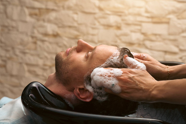 Ο κομμωτής λούζει τα μαλλιά του πελάτη στο κουρείο. Άνδρας πελάτης κουρεύεται από κομμωτή. Φροντίδα μαλλιών, βιομηχανία ομορφιάς, κουρέας έννοια. - Φωτογραφία, εικόνα