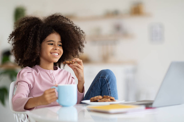 Cheerful black girl with bushy hair having break while studying at home, sitting at desk, eating cookies, drinking juice or cocoa, looking at computer screen and smiling, watching cartoons, copy space - Photo, Image