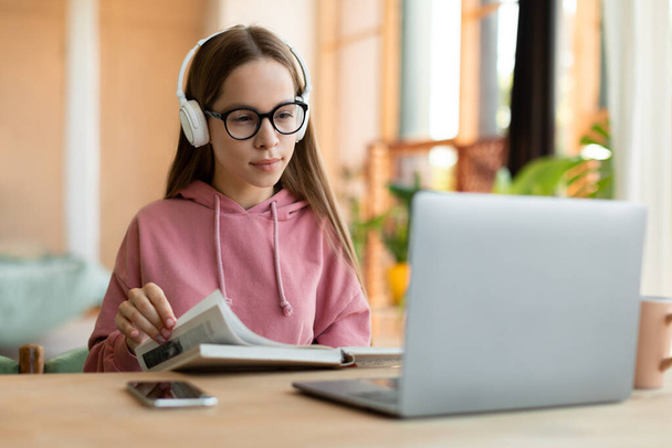 Focused female teenager studying, reading notebook and looking at laptop screen, learning remotely from home. Interested teen girl getting ready for online test or exam - Foto, imagen