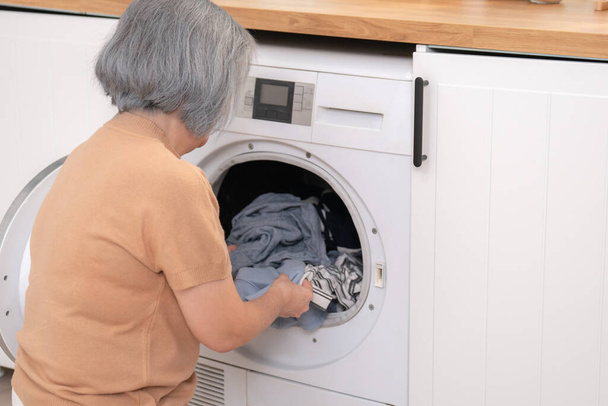 Задоволена старша домогосподарка займається пранням у пральні з одягом всередині пральної машини. Домашнє життя, сушильна машина, домашні завдання
. - Фото, зображення