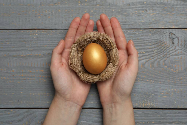 Woman holding nest with shiny golden egg at grey wooden table, top view - Φωτογραφία, εικόνα