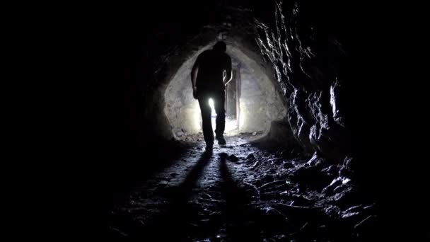 Egy fickó zseblámpával kijön egy sötét barlangból. A fény az alagút végén van. Sziklás falak és mennyezet. Egy mesterséges alagút a hegyekben. Régi rozsdás ajtók. Egy férfi árnyéka. - Felvétel, videó