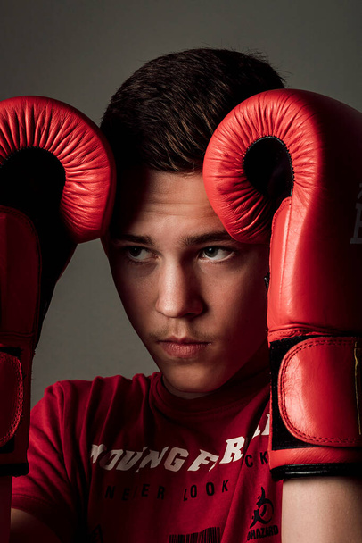 The guy pressed the gloves to his head and looked to the side with a frown. The guy is wearing a red T-shirt and red gloves. Dark background behind - Foto, Bild