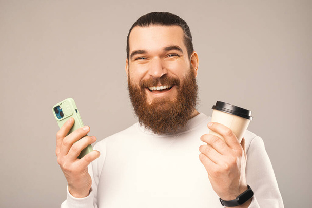 Man wide smiling at the camera is holding a take away coffee cup and a phone. Close up studio portrait over warm grey background. - Foto, afbeelding