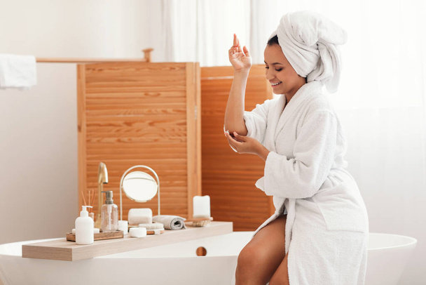 Skincare. Cheerful Woman Applying Moisturizer Cream On Elbow Moisturizing Skin Sitting On Bathtub In Bathroom At Home. Bodycare Cosmetics And Beauty Routine, Wellness And Spa Concept - Fotó, kép