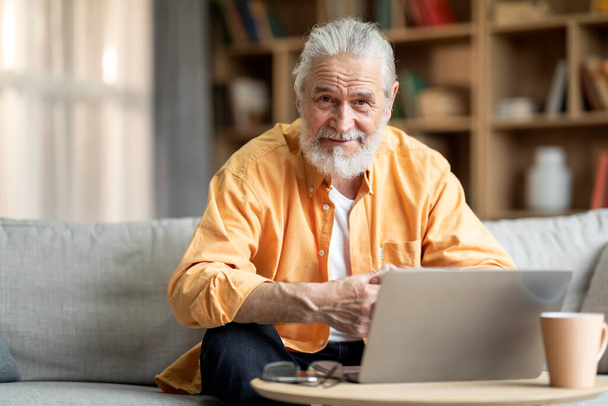 Handsome bearded grey-haired senior man freelancer working from home, sitting on couch, using laptop, drinking coffee, looking at camera and smiling, copy space. Job opportunities for elderly people - 写真・画像