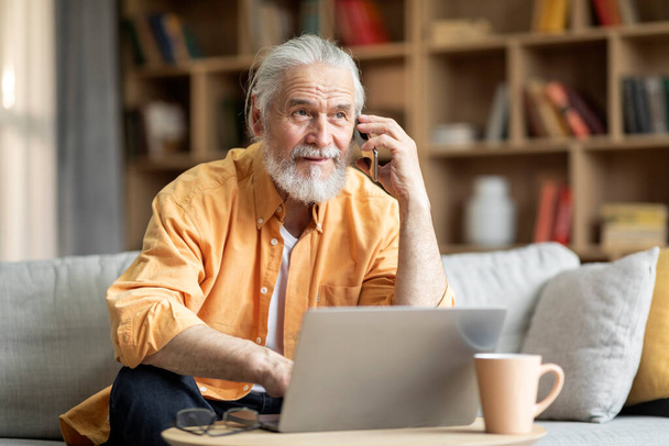 Cheerful handsome elderly man with beard businessman sitting on couch, using modern laptop and having phone conversation with assistant, working from home, looking at copy space - Photo, Image