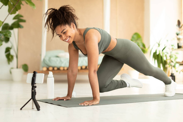 Online workout. Fit african american female doing cross body mountain climbers exercise on yoga mat, looking at smartphone on tripod, recording video for blog or having chat with coach - 写真・画像