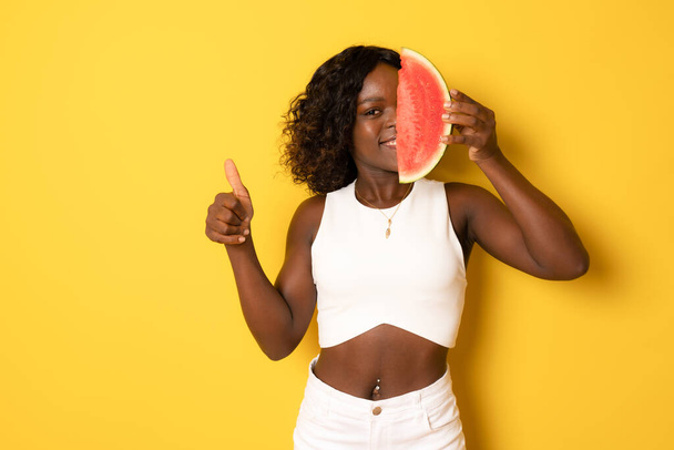 Photo of excited dark skin girl bite natural watermelon slice wear white t-shirt showing thumb up isolated on yellow color background - Fotó, kép