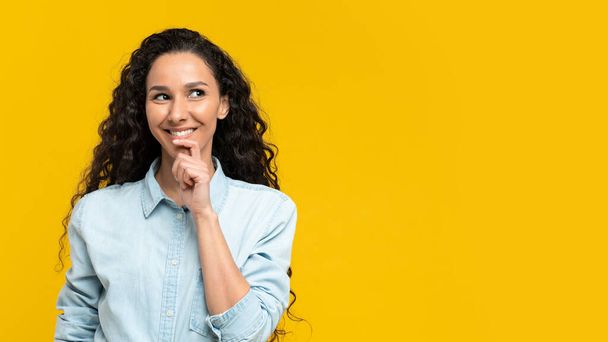Happy thoughtful young woman in casual outfit looking at empty space over yellow studio background, banner design. Positive millennial female offering free place for ad or promo - Zdjęcie, obraz