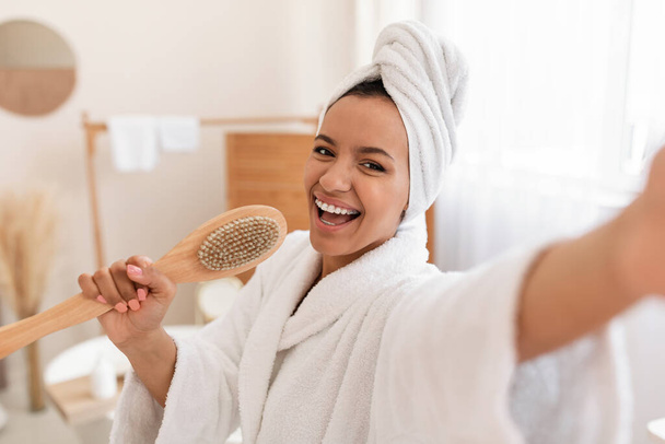 Cheerful Woman Having Fun Caring For Body Singing Holding Wooden Massage Brush Like Microphone Standing In Modern Bathroom Indoor, Posing With Towel On Head. Beauty And Bodycare Cosmetics - 写真・画像