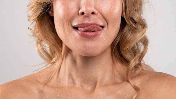 Cropped Image Of Beautiful Middle Aged Female Playfully Licking Lips With Tongue, Closeup Shot Of Unrecognizable Blonde Woman With Bare Shoulders Standing Over Light Grey Studio Background - Photo, image