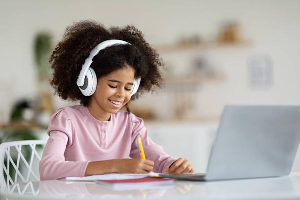 Cheerful african american schooler cute preteen girl with bushy hair sitting at table in front of laptop, using wireless headset, taking notes, doing homework at home, copy space - Photo, Image