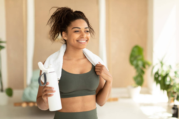 Portrait of happy black woman in sportswear and white towel on neck holding bottle with fresh water, having rest after domestic workout, free copy space - Valokuva, kuva