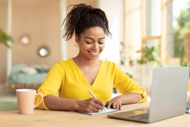 Note taking concept. Happy young black woman using pc laptop and writing in her workbook, millennial female holding pen preparing short summary for educational project, sitting at desk - Фото, изображение