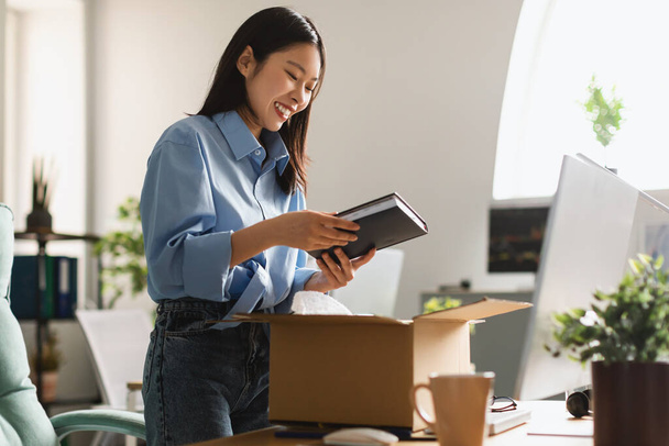 Cheerful Asian Businesswoman Unpacking Box Holding Book Standing In Modern Office Indoors. Business Literature, Shopping And Delivery Service Concept. Side View - Photo, Image
