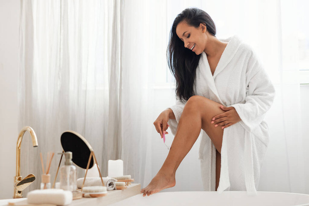 Happy Female Shaving Legs Removing Hair With Safety Razor Wearing Bathrobe Standing In Modern Bathroom Indoor. Hair Removal, Depilation And Body Care Routine Concept - Φωτογραφία, εικόνα