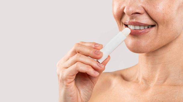 Lips Care. Smiling middle aged woman applying lip balm, closeup of unrecognizable mature female using hygienic lipstick, lady with bare shoulders standing over light studio background, copy space - Foto, Bild