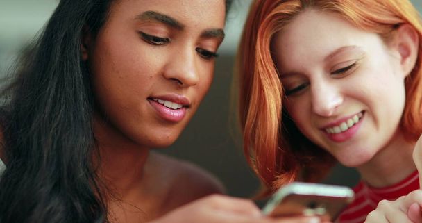 Two multi-cultural girls looking at cellphone screen, ethnically diverse young women holding smartphone device - Fotó, kép