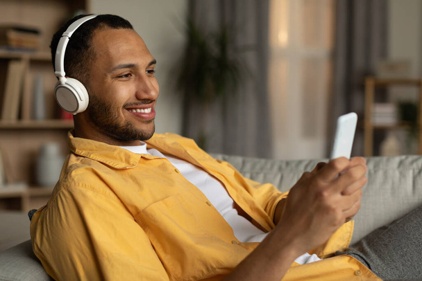 Joyful young black guy in headphones lying on couch and listening to music online on cellphone at home. Cool millennial African American man watching video on smartphone, using wireless headset - Foto, immagini