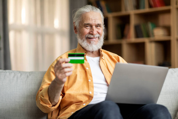 Smiling bearded senior man sitting on couch, using modern laptop and credit card, shopping on Internet while staying at home, paying for goods and sevices online, copy space - Foto, immagini