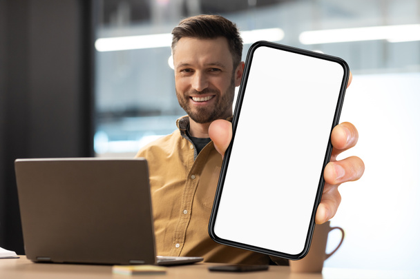 Business Offer. Smiling Millennial Man Demonstrating Big Blank Smartphone While Sitting At Desk With Laptop In Office, Cheerful Young Businessman Recommending Mobile App Or Website, Collage, Mockup - 写真・画像