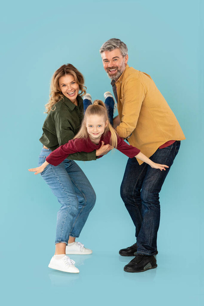 Cheerful Parents Playing With Little Daughter Holding Her In Arms Posing On Blue Background. Studio Shot Of Family Having Fun Together, Kid Spreading Hands Like Plane. Vertical - Фото, изображение
