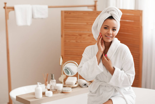 Attractive Lady Touching Face Looking At Camera Posing Caring For Herself Wearing White Bathrobe Sitting In Modern Bathroom At Home. Beauty And Facial Skincare Cosmetics Concept - Foto, Bild