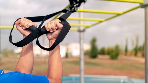 Close up hands of young man doing fitness exercises on a special hanging device. Fitness male guy doing exercising with suspension trainer slingin a city park. A suspended TRX sport fitness concept. - Foto, Imagen