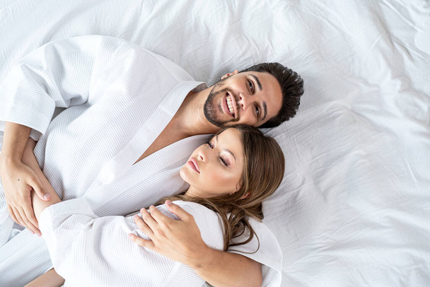 Young happy couple relaxing, lying in bed in white bathrobes, hugging and smiling. Morning light. Real people lifestyle. - Fotoğraf, Görsel