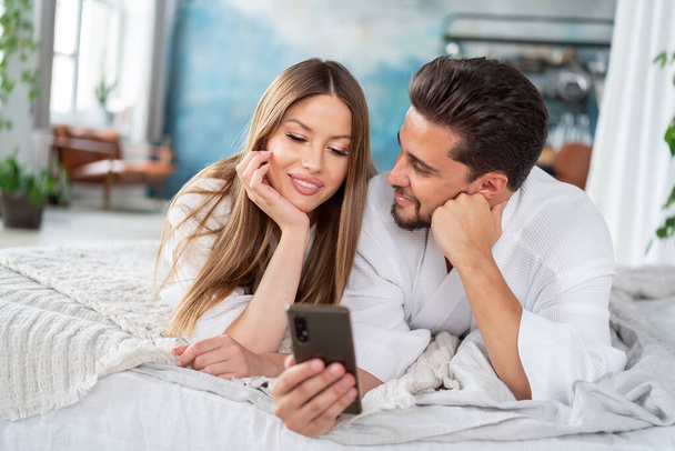 Happy young couple relaxing on bed at home in bathrobes, using mobile phone, taking selfie or scrolling social media together, having fun. Real people lifestyle, emotions. Date. - Photo, image
