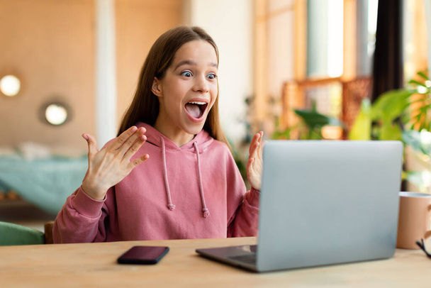 Нічого собі. Здивована дівчина-підліток використовує ноутбук, дивлячись на екран з відкритим ротом з подивом, читаючи новини або отримуючи A-клас, сидячи за столом вдома
 - Фото, зображення