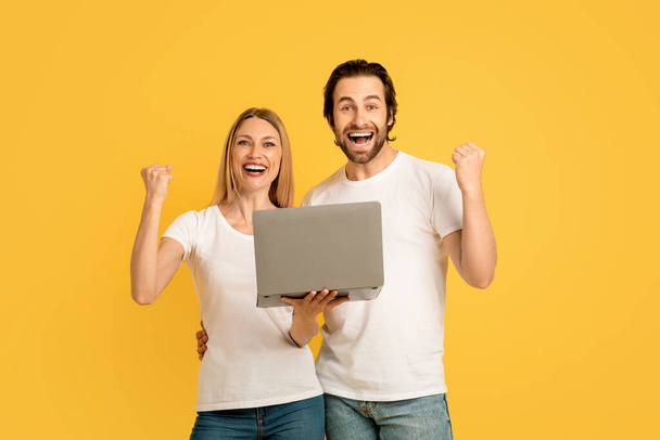 Smiling glad millennial caucasian guy and lady in white t-shirts hold laptop, making victory sign with hands, isolated on yellow background. Success, online win with technology, great offer and sale - Photo, image