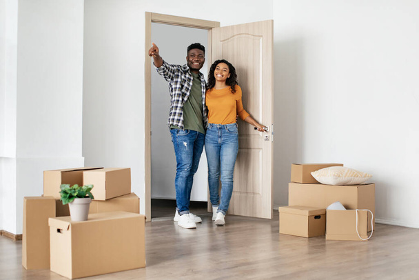 Real Estate Ownership. African American Husband And Wife Entering Their New House Standing In Living Room Among Moving Boxes At Home. Apartment Rent And Purchase. Full Length Shot - Foto, Imagen