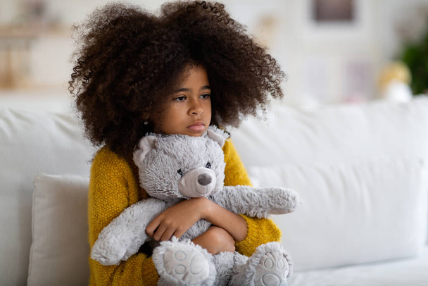 Upset preteen african american girl with curly bushy hair sitting on couch alone, embracing teddy bear, looking at copy space. Kid feeling lonely at home, missing parents or friends - Photo, image