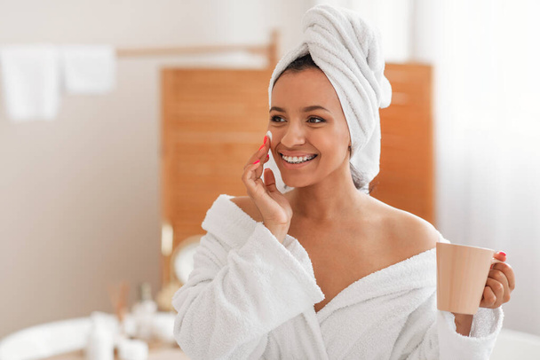 Facial Skincare. Happy Young Female Applying Moisturizer On Face Moisturizing Skin Holding Coffee Cup Standing Smiling Looking Aside In Modern Bathroom Indoors. Beauty And Cosmetics - Φωτογραφία, εικόνα