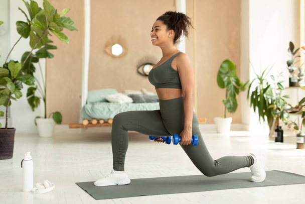 Strength training concept. Sporty african american woman doing lunges exercise with dumbbells, working out on yoga mat at home, full length. Black lady leading healthy lifestyle - Photo, image