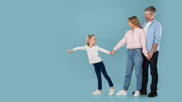 Family Offer. Little Daughter Pointing Finger Aside At Copy Space Posing Holding Hands With Middle Aged Parents Standing Over Blue Studio Background. Full Length, Panorama - Фото, изображение