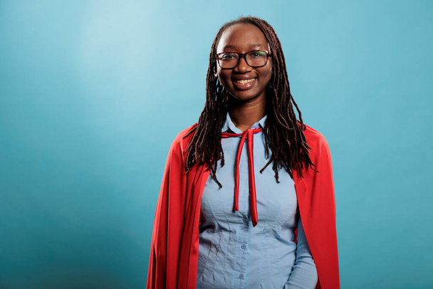 Giovane affascinante bella sorridente donna supereroina africana americana indossa mantello rosso su sfondo blu. Ambizioso adulto ottimista guardando orgoglioso in macchina fotografica pur essendo felice e gioioso. - Foto, immagini