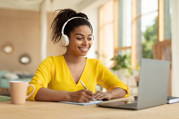 Smiling black lady in wireless headphones working on laptop and taking notes in paper notebook, watching webinar, sitting at desk at home, free copy space - Фото, изображение