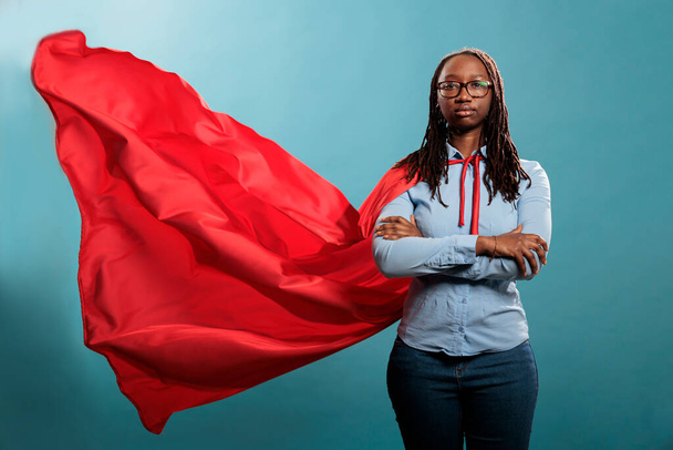 Confident and brave young superhero woman standing with arms crossed while wearing red mighty cape on blue background. Portrait of justice defender posing tough and strong while looking at camera. - Fotó, kép