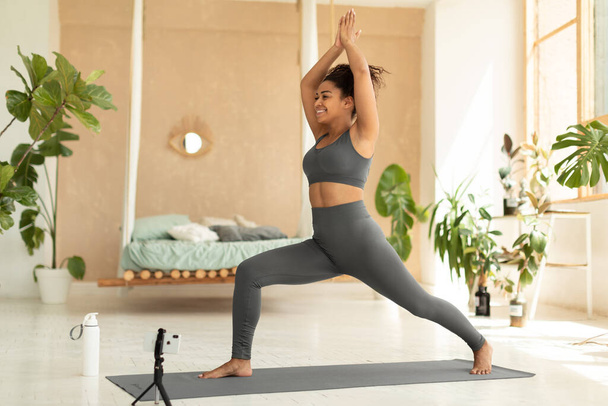 Young african american lady standing in 1 warrior pose on yoga mat, shooting online workout video tutorial on cellphone, doing forward lunges exercise with arms overhead - Фото, изображение