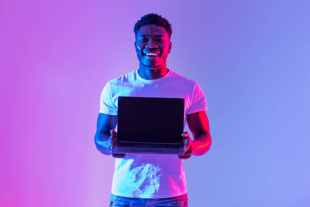 Check this website. Handsome young black man holding laptop with blank screen in neon light, mockup for design. Happy African American male showing empty space for online ad - Foto, immagini