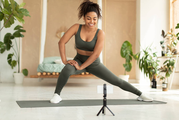 Happy fit black lady in sportswear doing side lunge exercise, stretching during home workout, training and looking at smartphone on tripod, making video call with coach - Photo, image