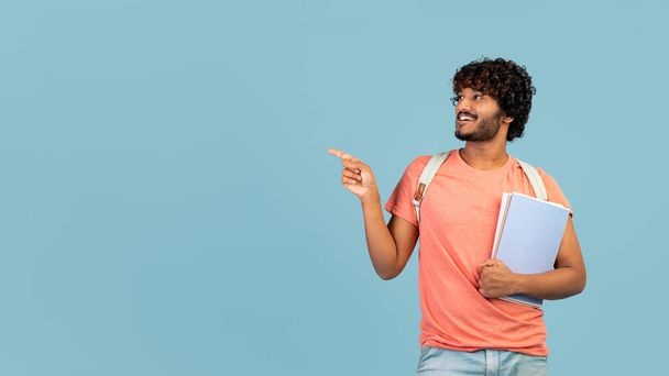 Happy young hindu guy going to college, carrying books, backpack, pointing at copy space and smiling over blue studio background, panorama. University, school, education concept - Φωτογραφία, εικόνα