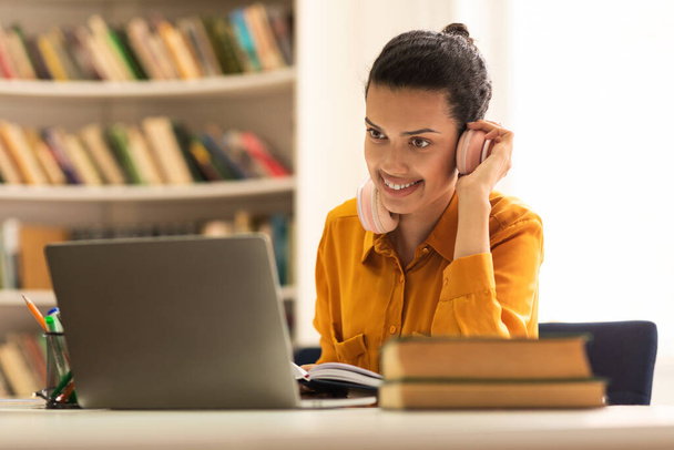 Happy mixed race lady touching wireless headphones and looking at laptop screen, sitting at desk in library, having webinar, studying online - Foto, Bild