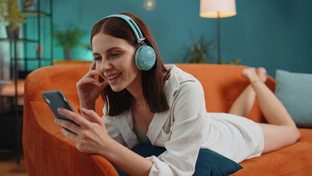 Молода жінка в бездротових навушниках розслабляється лежачи на дивані вдома, вибираючи слухати улюблену енергійну диско-рок-н-ролу танцювальну музику в смартфоні. Люди у вихідні дні. Щаслива насолоджена дівчина
 - Фото, зображення