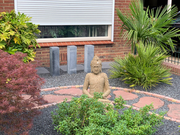 Beautiful stone statue with a green leaves. Oriental style house entrance with cement Buddha statue and stone garden path and oriental plants: buxus, acer japonicum, fatsia japonica and palm - Foto, afbeelding