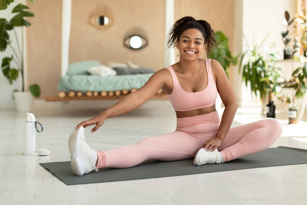 Domestic sports concept. Happy fit black lady in sportswear doing fitness exercises on yoga mat, stretching her legs and smiling at camera. Sporty woman staying flexible, working out at home - Foto, imagen